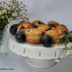 Pyszne muffinki ze śliwkami, świeże i pachnące, ułożone na białej paterze. Obok bukiet drobnych kwiatów i dojrzałe śliwki.