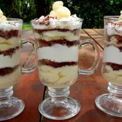 Trzy pucharki z deserkiem z mascarpone, przełożonymi kremem i czekoladowymi okruszkami, ozdobione bitą śmietaną i startą czekoladą.