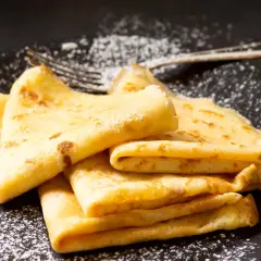 Puszyste naleśniki, delikatnie posypane cukrem pudrem, czekają na talerzu. Obok leży widelec, gotowy do degustacji.