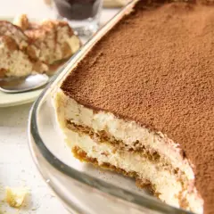 Pyszne tiramisu przepis w naczyniu, posypane kakao. Obok porcja na talerzyku i biszkopty.