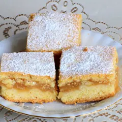 Trzy kawałki puszystego ciasta z jabłkami, posypane cukrem pudrem, na białym talerzyku.
