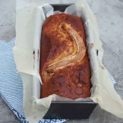 Szybkie ciasto bananowe z olejem, świeżo upieczone, w keksówce wyłożonej papierem, z bananem na wierzchu.
