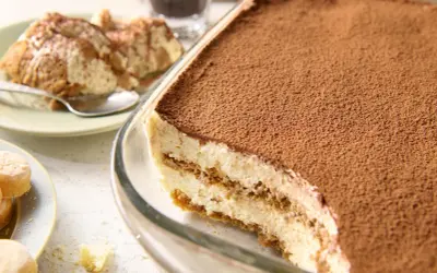 Pyszne tiramisu przepis w naczyniu, posypane kakao. Obok porcja na talerzyku i biszkopty.