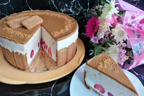 Pyszny sernik lotus z truskawkami, ozdobiony ciasteczkami i posypką. Obok bukiet kwiatów.