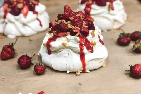 Słodkie mini pavlova z bitą śmietaną, truskawkami i sosem truskawkowym, posypane kruszonką.