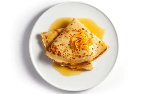 Złożone na pół naleśniki crepes suzette polane pomarańczowym sosem, ozdobione skórką pomarańczową.