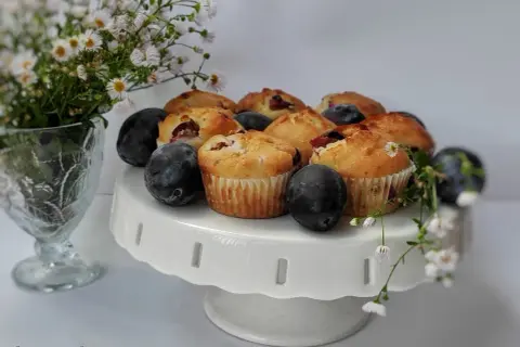 Pyszne muffinki ze śliwkami, świeże i pachnące, ułożone na białej paterze. Obok bukiet drobnych kwiatów i dojrzałe śliwki.