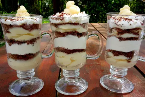 Trzy pucharki z deserkiem z mascarpone, przełożonymi kremem i czekoladowymi okruszkami, ozdobione bitą śmietaną i startą czekoladą.