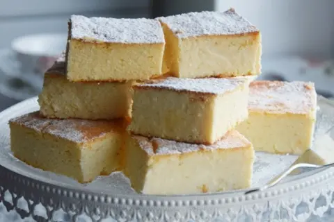 Stos kawałków tradycyjnego sernika, posypanych cukrem pudrem, ułożonych na paterze. Idealny na deser.
