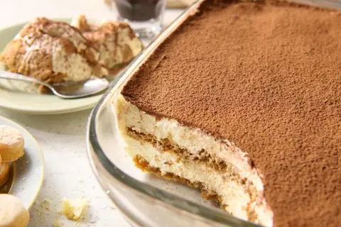 Pyszne tiramisu przepis w naczyniu, posypane kakao. Obok porcja na talerzyku i biszkopty.