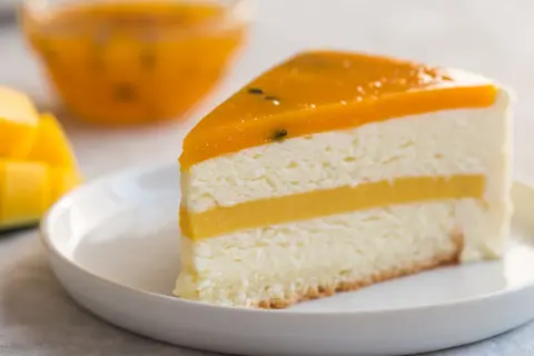 Pyszny tort mango z warstwami musu i galaretki, podany na białym talerzyku. Obok kawałki świeżego mango.