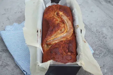 Szybkie ciasto bananowe z olejem, świeżo upieczone, w keksówce wyłożonej papierem, z bananem na wierzchu.