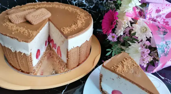 Pyszny sernik lotus z truskawkami, ozdobiony ciasteczkami i posypką. Obok bukiet kwiatów.