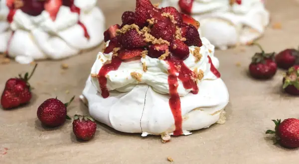 Słodkie mini pavlova z bitą śmietaną, truskawkami i sosem truskawkowym, posypane kruszonką.