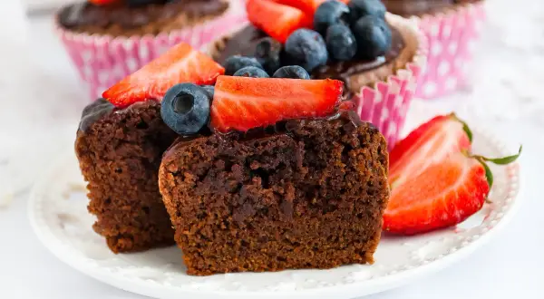 Pyszne babeczki brownie z polewą czekoladową, udekorowane truskawkami i borówkami, kuszą swoim wyglądem.