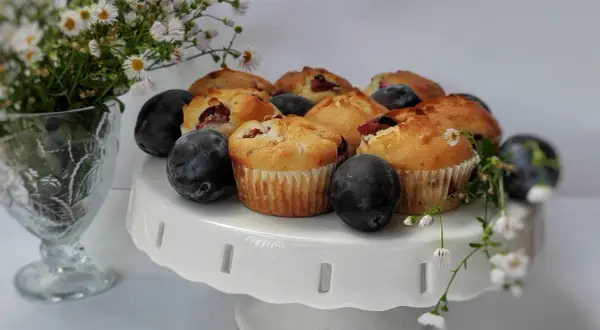 Pyszne muffinki ze śliwkami, świeże i pachnące, ułożone na białej paterze. Obok bukiet drobnych kwiatów i dojrzałe śliwki.