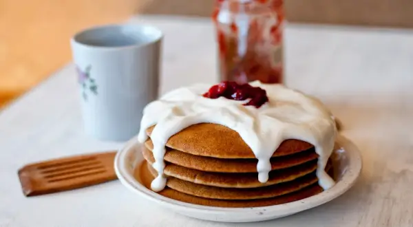 Pyszne pancakes bezglutenowe polane kremowym jogurtem i dżemem, obok kubek i słoiczek z konfiturą.