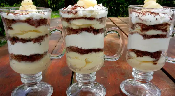 Trzy pucharki z deserkiem z mascarpone, przełożonymi kremem i czekoladowymi okruszkami, ozdobione bitą śmietaną i startą czekoladą.