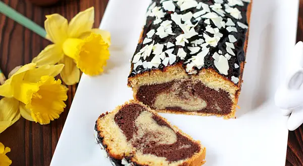 Wilgotna babka marmurkowa z polewą czekoladową i płatkami migdałów, ozdobiona żółtymi żonkilami.