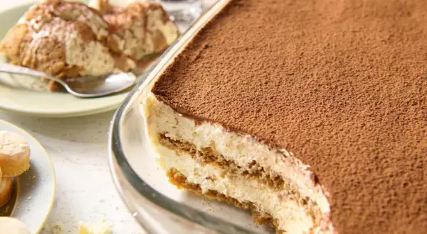 Pyszne tiramisu przepis w naczyniu, posypane kakao. Obok porcja na talerzyku i biszkopty.