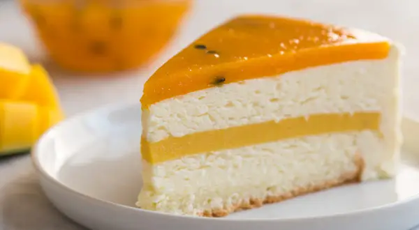 Pyszny tort mango z warstwami musu i galaretki, podany na białym talerzyku. Obok kawałki świeżego mango.