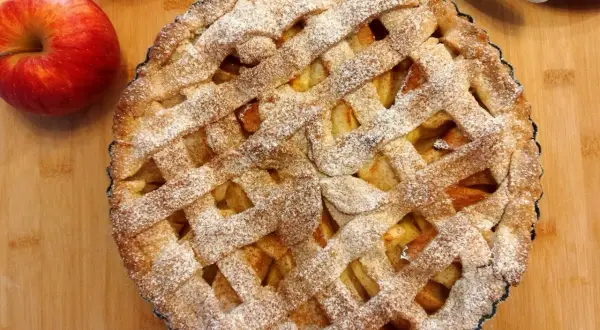 Pyszna tarta z jabłkami, posypana cukrem pudrem, leży na drewnianej desce obok świeżych jabłek.