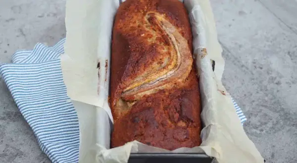 Szybkie ciasto bananowe z olejem, świeżo upieczone, w keksówce wyłożonej papierem, z bananem na wierzchu.