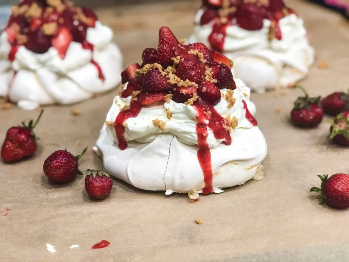 Słodkie mini pavlova z bitą śmietaną, truskawkami i sosem truskawkowym, posypane kruszonką.