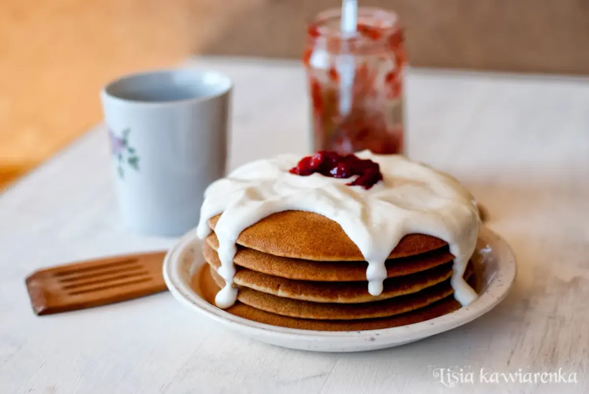Pyszne pancakes bezglutenowe polane kremowym jogurtem i dżemem, obok kubek i słoiczek z konfiturą.