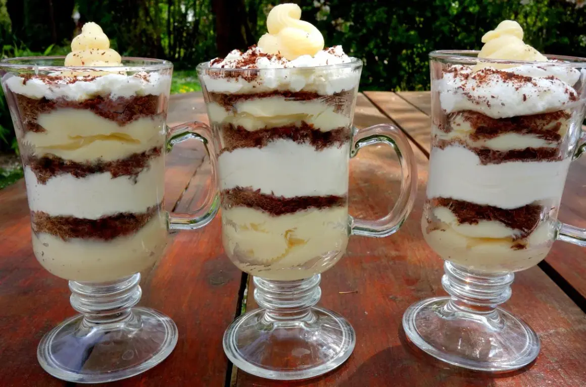 Trzy pucharki z deserkiem z mascarpone, przełożonymi kremem i czekoladowymi okruszkami, ozdobione bitą śmietaną i startą czekoladą.