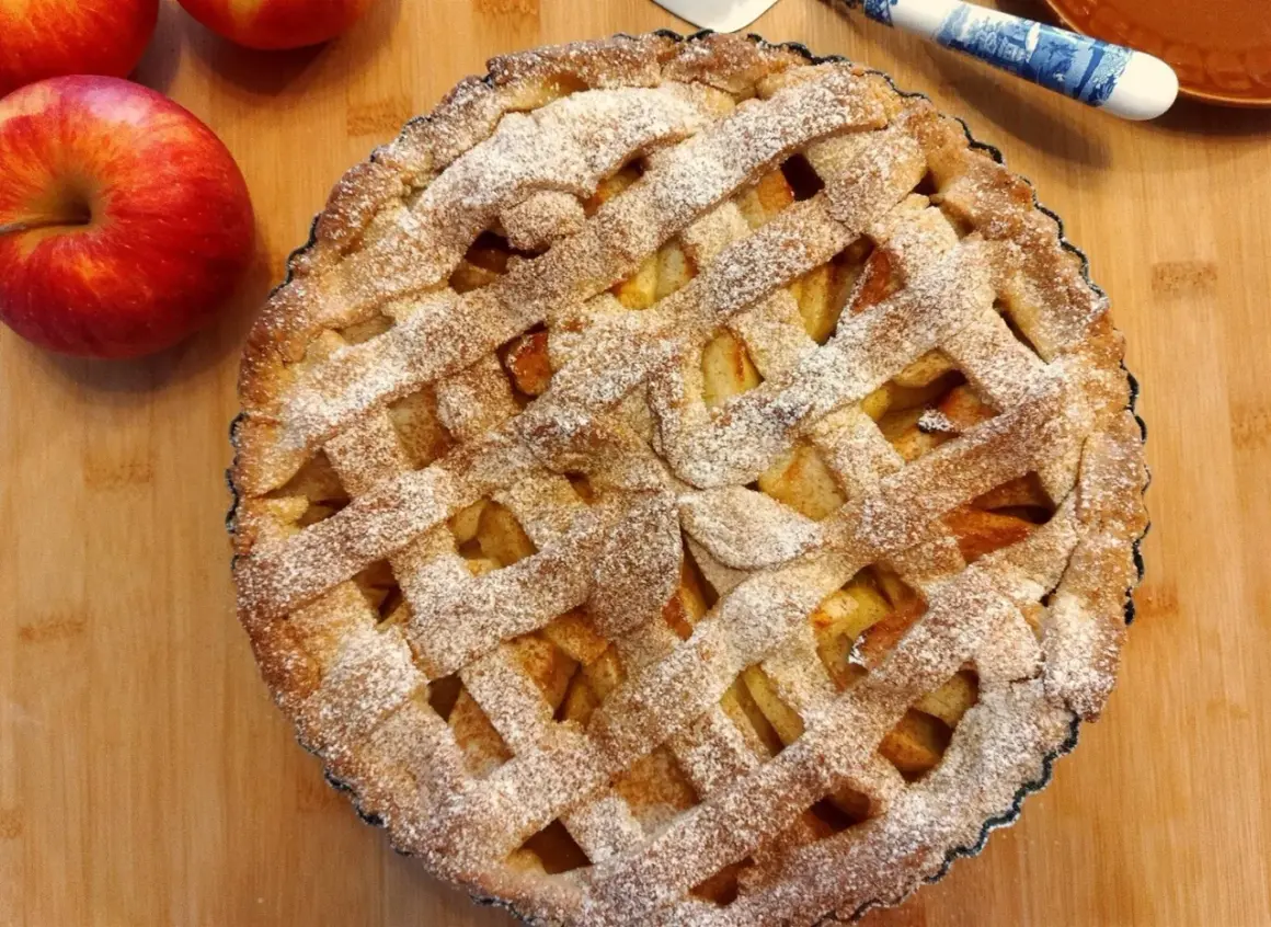 Pyszna tarta z jabłkami, posypana cukrem pudrem, leży na drewnianej desce obok świeżych jabłek.