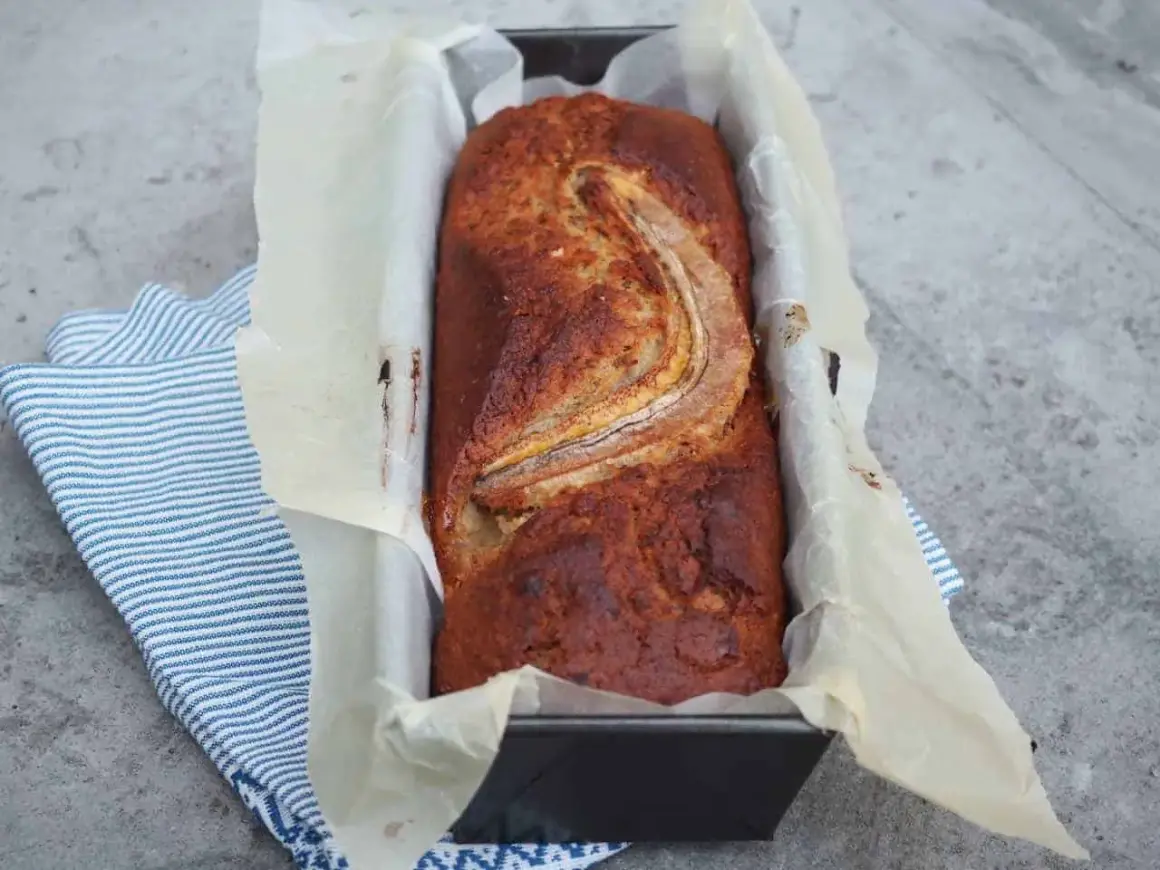 Szybkie ciasto bananowe z olejem, świeżo upieczone, w keksówce wyłożonej papierem, z bananem na wierzchu.