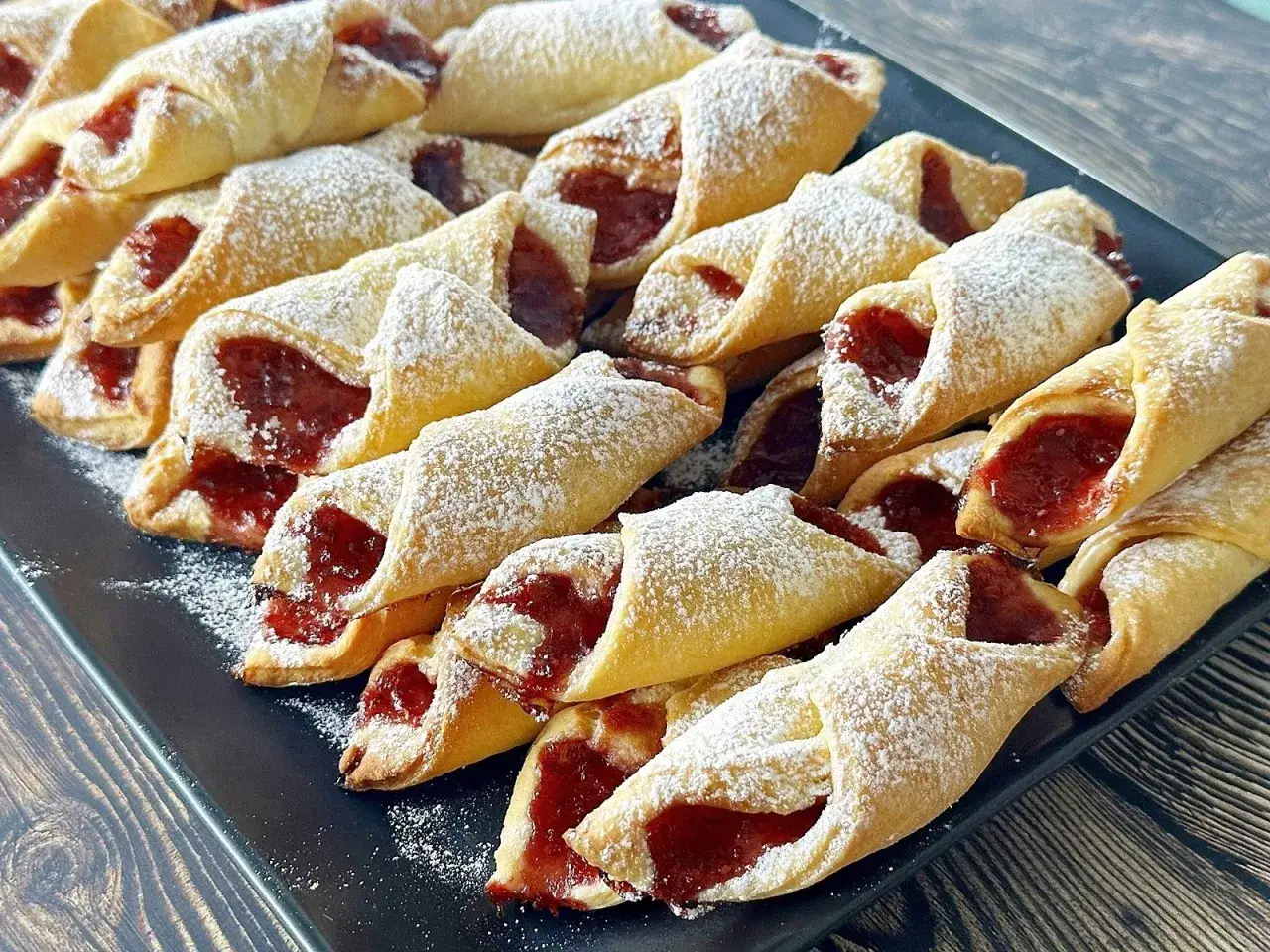 Szybkie kruche ciasteczka z dżemem, posypane cukrem pudrem, ułożone na czarnym talerzu.