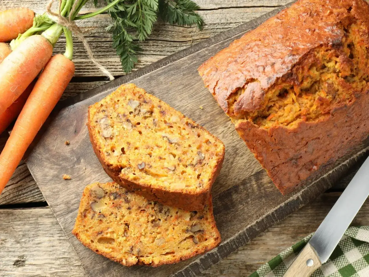 Wilgotne ciasto marchewkowe z orzechami, pokrojone na kromki, leży na desce obok świeżych marchewek.