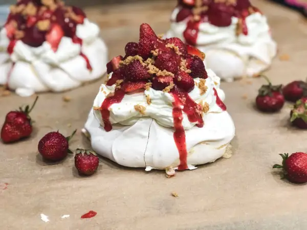 Słodkie mini pavlova z bitą śmietaną, truskawkami i sosem truskawkowym, posypane kruszonką.