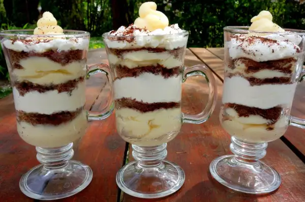 Trzy pucharki z deserkiem z mascarpone, przełożonymi kremem i czekoladowymi okruszkami, ozdobione bitą śmietaną i startą czekoladą.